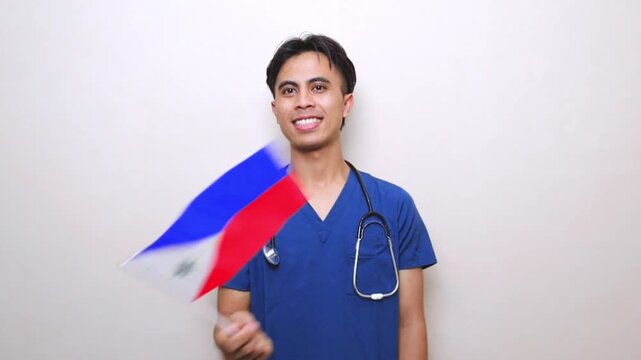 Joyful Filipino male nurse in blue scrubs holding and waving a small Philippines national flag, celebrating Independence Day or medical achievement