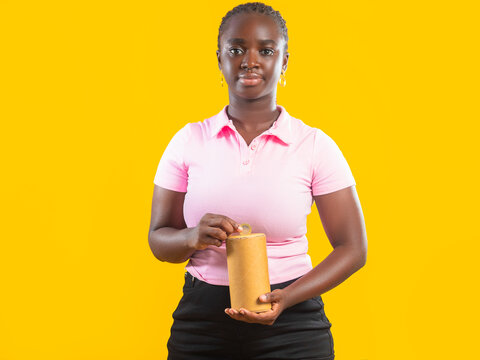 Close-up of a Black woman putting a coin into a cardboard savings box against a yellow background. Represents financial planning, budgeting, micro-investing, and personal savings goals.