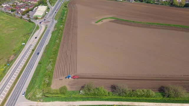 Aerial view of a blue tractor plowing a large brown field next to a busy road and green trees in Knottingley, England, United Kingdom.
