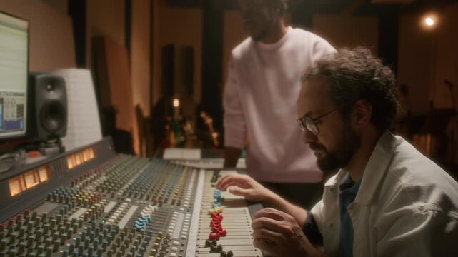Professional sound engineer adjusting faders on large mixing console and looking at digital audio workstation on monitor in recording studio, Black music artist standing beside and observing process