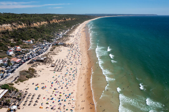 Aerial drone shot of Praia de Fonte da Telha beach in Costa de Caparica, Portugal. Sunbathers and swimmers enjoy the packed beach while the parking lot fills