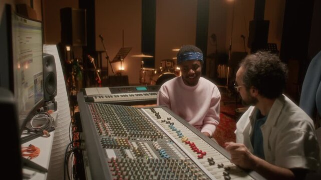 Young Black musician and Caucasian sound engineer sitting by mixing desk, smiling and talking as working together during audio mastering session in professional recording studio