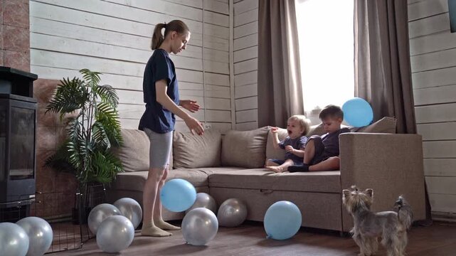 Teen babysitter tossing blue balloons gently, two kids reclining on sofa reach and giggle while small terrier watches, afternoon window light and scattered metallic balloons set calm playful mood