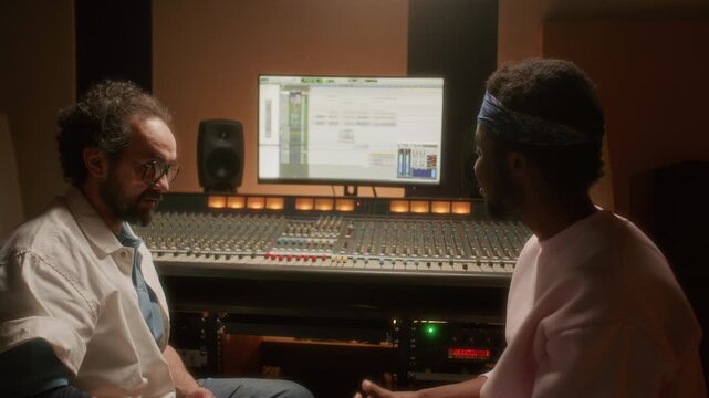Young Black musician explaining his creative ideas to Caucasian sound engineer, he listening attentively and adjusting faders on mixing console while mastering audio tracks in recording studio