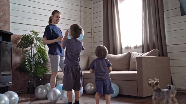 Children playing with blue balloons indoors, siblings and teen caregiver toss balloons near sofa and plant while small dog jumps, warm natural light and scattered silver balloons create intimate