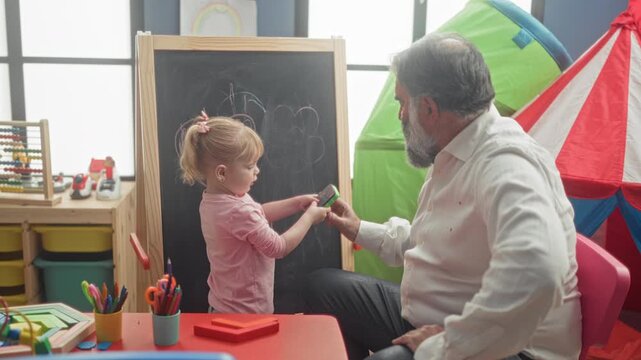 Man erases chalk on a chalkboard while guiding a little girl who draws a heart at a table in a children's play area inside a building; learning bond warmth.