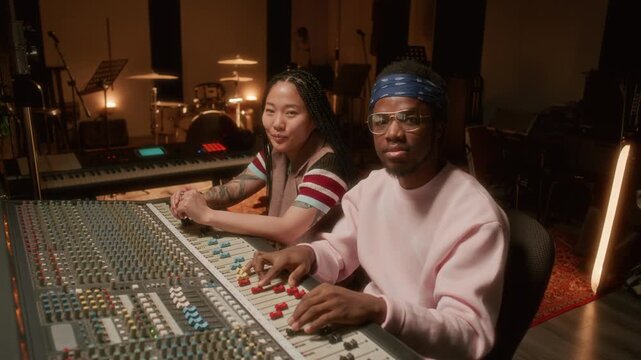 Portrait of young Asian female musician and Black male sound engineer sitting by mixing console, looking at camera and smiling while working together in audio recording studio
