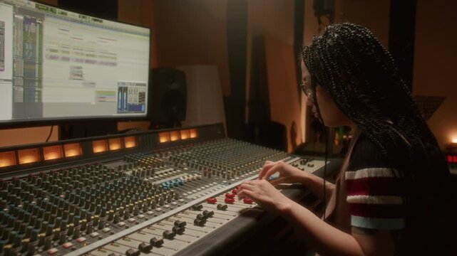 Asian female sound engineer with braided hair and tattoos operating mixing console and monitoring digital audio workstation in professional recording studio