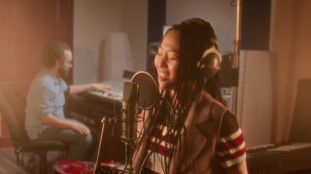 Young Asian female vocalist in headphones singing emotionally into professional condenser microphone with pop filter, audio engineer working at mixing console in background during recording session