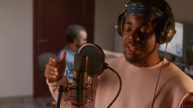 Young Black musician wearing blue bandana and headphones singing emotionally into professional condenser microphone in studio, sound engineer monitoring recording session at workstation in background