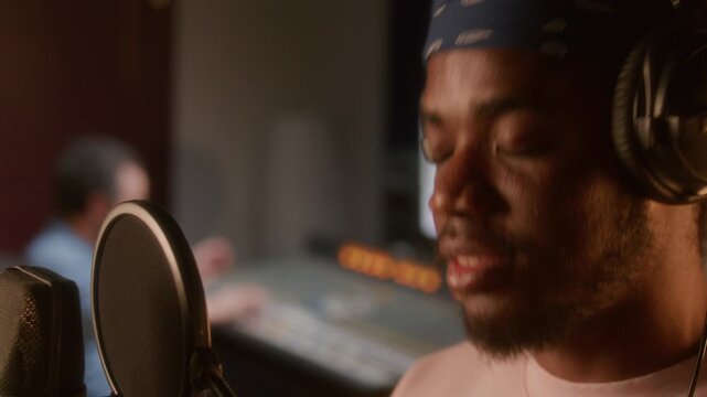Young soulful African American singer in headphones performing into professional condenser microphone with pop filter in recording studio, sound engineer working in background. Close up view