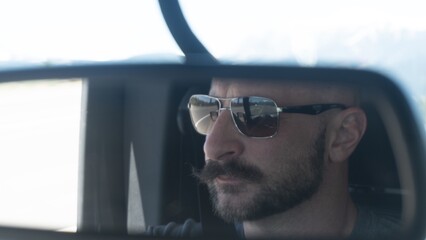 A cinematic close-up shot focused on the rearview mirror of a vehicle, capturing the reflection of a middle-aged man with a prominent mustache and dark aviator-style sunglasses. The driver appears foc © Christopher