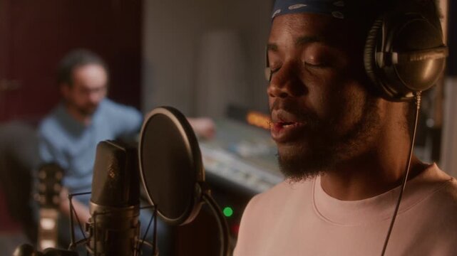 Young Black male vocalist with bandana and headphones singing soulfully into microphone in professional audio studio while recording song with music producer working at mixing desk in background