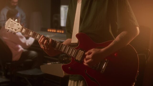 Close up midsection shot of musician playing red electric guitar in warmly lit recording studio, producer and sound engineer working at mixing console in background