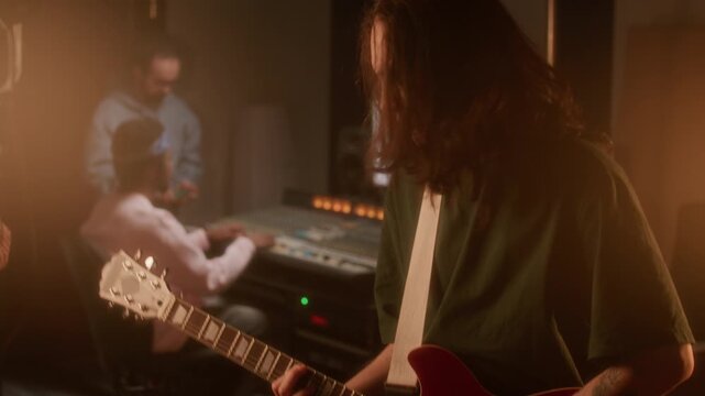 Emotional young musician with long hair and tattoos playing electric guitar during recording session in audio studio bathed in warm light, sound technicians working together in background