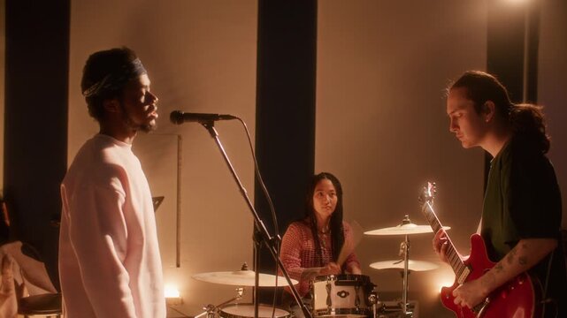 Multiethnic music band rehearsing in warmly lit studio: Black male vocalist singing into microphone, Caucasian guitarist playing electric guitar and Asian female drummer performing in background
