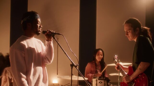 Diverse indie rock band rehearsing together in studio: African American vocalist singing into microphone, Asian woman drumming and Caucasian musician playing electric guitar under warm light