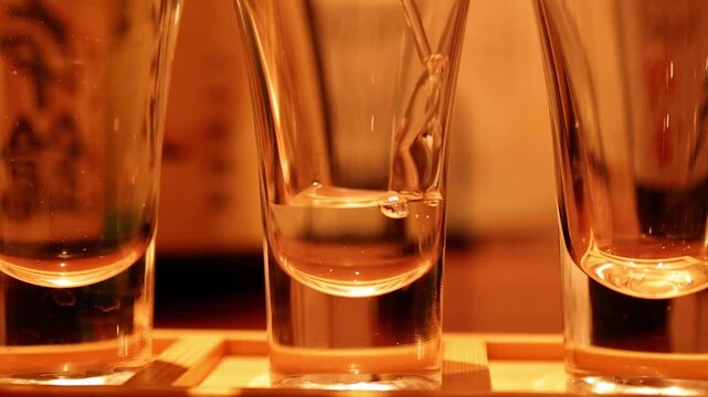 Clear liquid pouring into small shot glass in slow motion creating dynamic ripples and bubbles on warm background.
