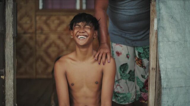 Mother placing a hand on her shirtless son's shoulder, both laughing inside their home