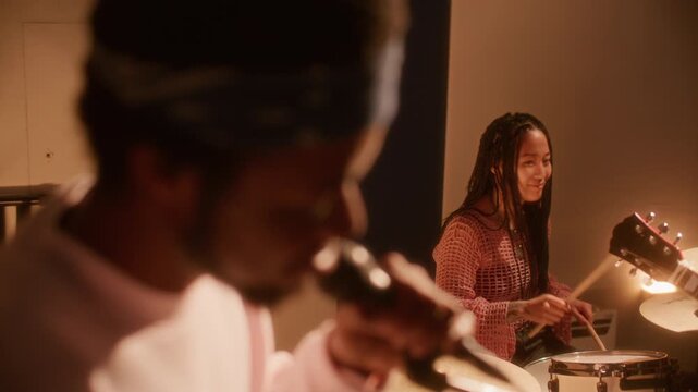 Multiethnic band performing together during studio session: young Asian woman playing acoustic drum kit as Black male vocalist holding microphone and singing under warm lighting. Rack focus