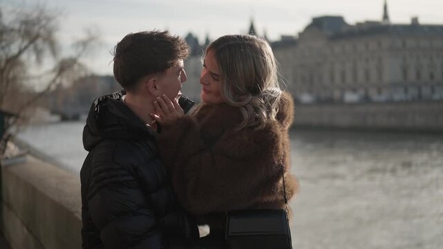 A romantic couple embraces along the Seine River in Paris France The woman wearing a fur coat lovingly holds the man's as they share a tender moment The city's iconic buildings form a picturesque back