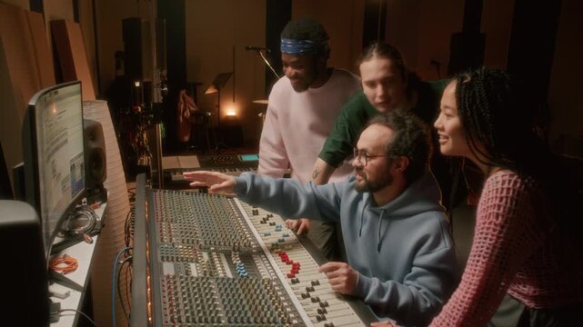 Music producer mastering audio tracks with young multiracial band in professional recording studio, operating mixing console and enjoying creative process