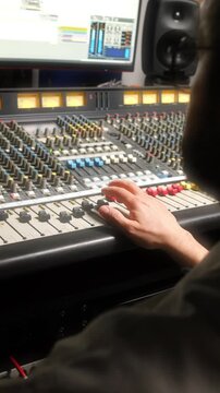 Professional sound engineer operating analog mixing desk in recording studio, adjusting channel faders and looking at computer monitor displaying music editing software. Vertical clip