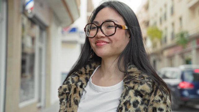 Woman, young latina, with glasses waving hand on a city street near storefronts, wearing leopard coat and smiling broadly; confidence.