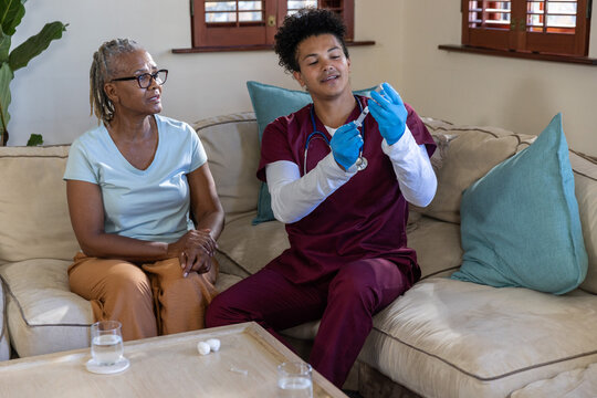 Senior African American woman watching mid adult man in maroon scrubs preparing syringe on sofa