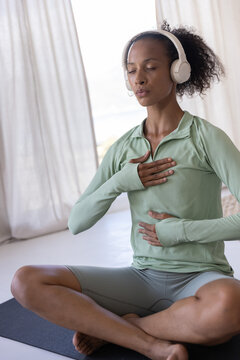 African American woman breathing deeply on yoga mat at home in sportswear with wireless headphones