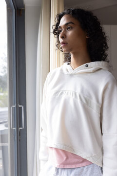 Nonbinary person wearing hoodie standing, gazing out sliding door inside with partly drawn curtain