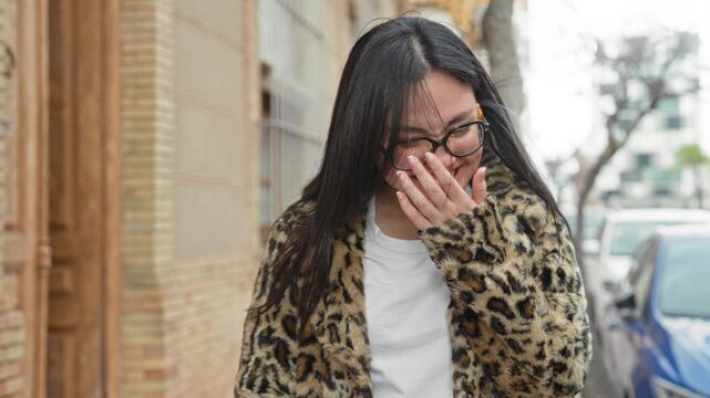 Woman smiling with hand near face, wearing round glasses and leopard print coat while walking past parked cars on a street; joy playfulness warmth.