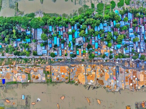 Kishoreganj, Bangladesh - 05 May 2026: Aerial view of a village settlement with blue-roofed houses, lush green trees, and agricultural grain drying areas by the water.