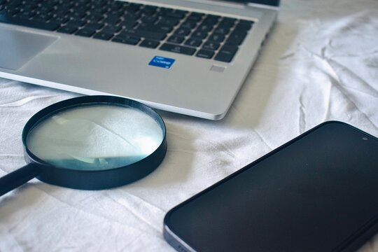 Sarawak, Malaysia - 18 April 2026: Still life of a magnifying glass, a smartphone, and a laptop on a white surface, showcasing the essential tools for modern research and digital investigation.