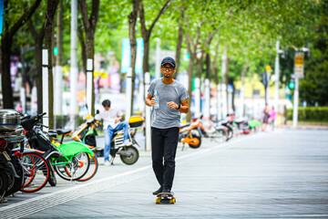 Asian man riding a skateboard and drinking water on a city sidewalk with parked scooters © Kenji