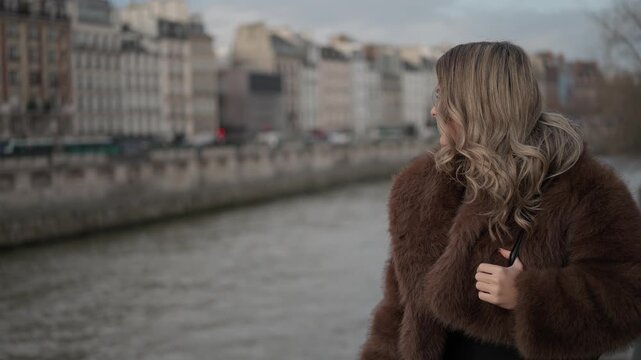 A woman in a fur coat gazes at the Seine River in Paris France The clip features a medium shot with a panning motion showcasing the romantic European scenery with Parisian architecture and the waterwa