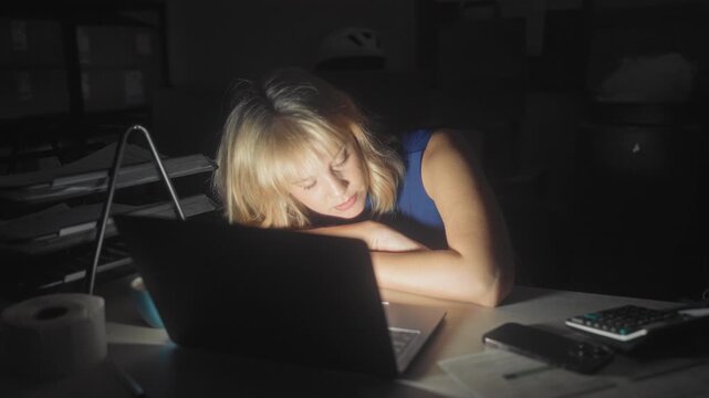 Woman rubbing eyes at laptop in a dim storage building with stacked parcels and a mug on the desk; late shift fatigue.