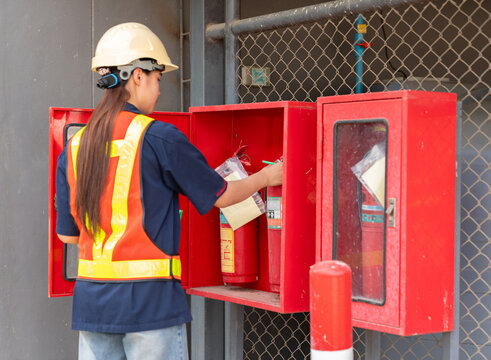 Female safety officer or inspector in a hard hat and reflective vest performing a maintenance check on fire extinguishers in a red storage cabinet at an industrial site.