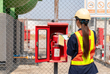 Female safety officer in a hard hat and reflective vest inspecting a fire extinguisher and recording the maintenance status on a checklist at an industrial facility. © Zblaster