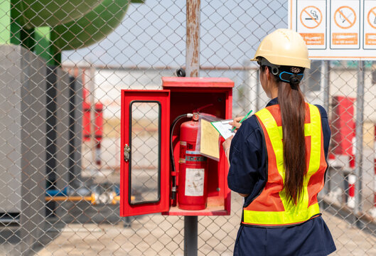 Female safety officer in a hard hat and reflective vest inspecting a fire extinguisher and recording the maintenance status on a checklist at an industrial facility.