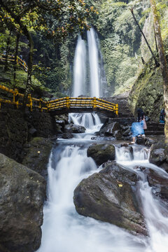 View of Grojogan Sewu waterfall with its powerful twin streams, a yellow wooden bridge, and mossy rocks surrounded by lush tropical forest in Kecamatan Tawangmangu, Jawa Tengah, Indonesia.
