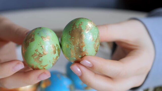 Two hands hold decorated green eggs with gold patterns. Eggs appear hand-painted for festive use. Background shows more colorful eggs out of focus. Hands gently rotate eggs slightly to show design
