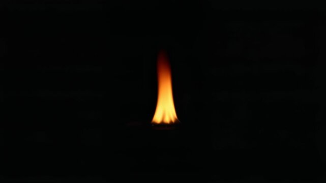 Small fire flame on dark background with minimal glow, warm orange light, isolated vertical shape, simple abstract focus, calm energy in black space