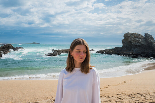 Young woman stand peaceful on a sandy beach looking down, with ocean rocks and blue sky background. Porto Covo, Setubal, Portugal