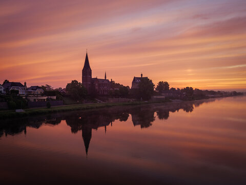 View of the silhouette of the historic church and village buildings reflected in the calm river Maas during a vibrant sunset in Kessel, Limburg, Netherlands.