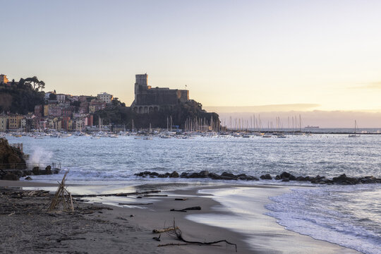 View of the historic Castello di Lerici overlooking the harbor filled with sailboats and colorful coastal houses from the sandy Venere Azzurra beach at sunset Lerici, Liguria, Italy.
