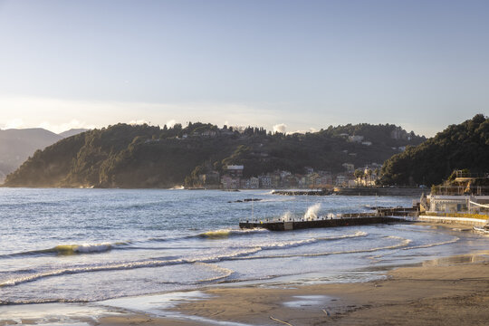 View of Venere Azzurra beach with waves crashing against a pier and lush green hills under a clear sky in Lerici, Liguria, Italy.