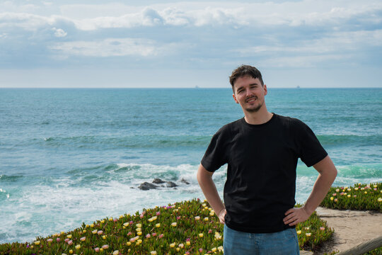 Confident Young Man Posing Against Dynamic Ocean Backdrop with Coastal Flowers on a Bright Day. Porto Covo, Setubal, Portugal