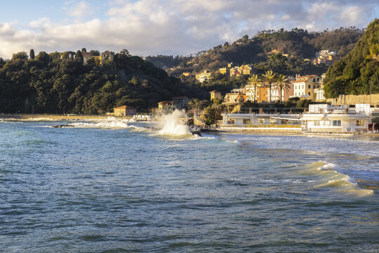 View of Venere Azzurra beach with waves crashing against the shore and colorful buildings on the hillside in Lerici, Liguria, Italy.
