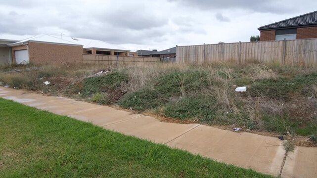 Overgrown vacant lot between family homes in a new residential area. Tarneit, Melbourne, Australia. The plot is covered weeds with scattered litter. Temporary wasteland in an outer suburb.